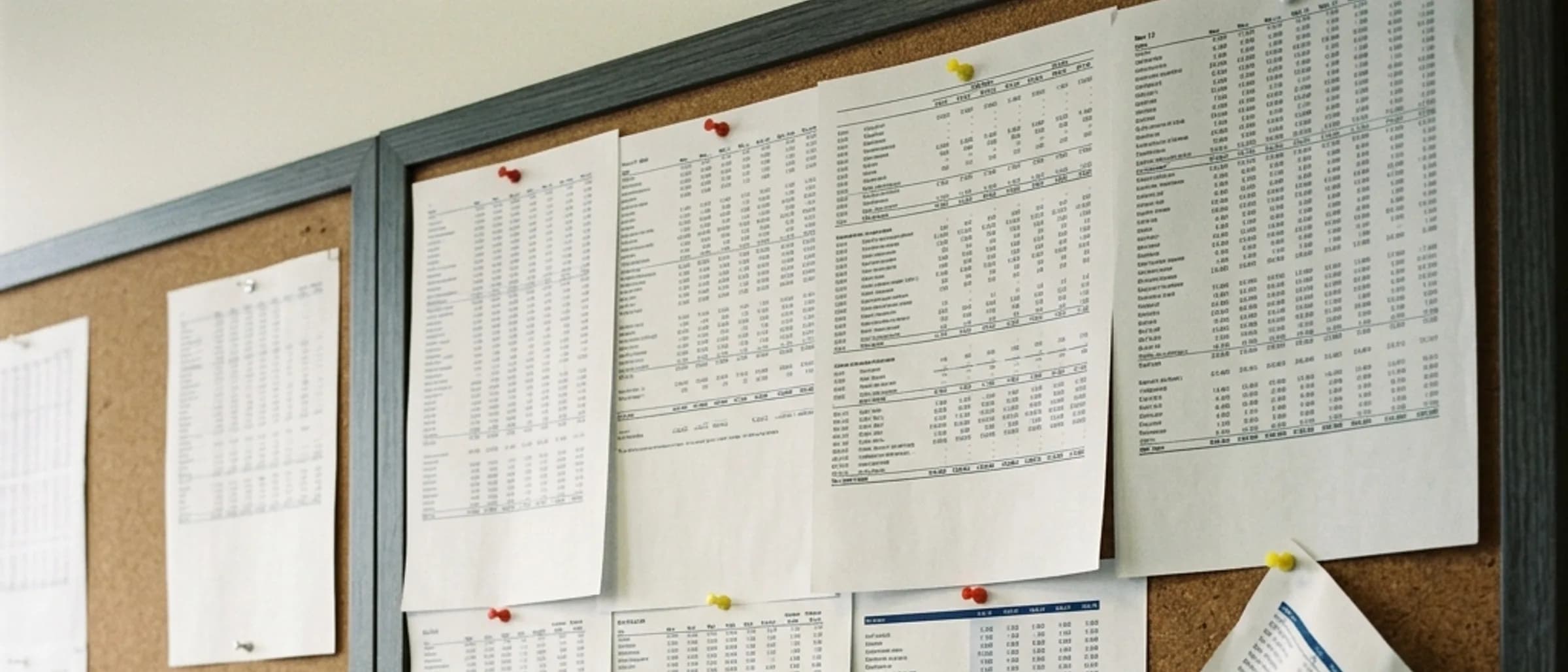 Printed spreadsheet pages pinned to a corkboard wall in an overlapping grid.